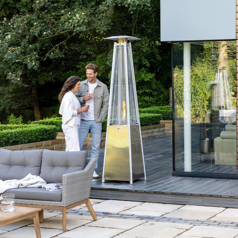 Tall metallic quadrilateral patio heater by Pacific Lifestyle, radiating warmth with its gas-fueled flames, positioned in a picturesque garden featuring lush grass and wooden decking. The background showcases a cozy outdoor setting with rattan seating, complemented by a man and woman engaged in conversation near the patio heater, creating a welcoming and intimate atmosphere during the day.