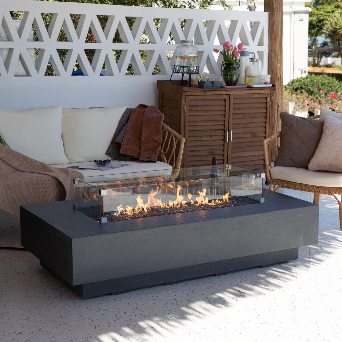 Long concrete grey fire pit table by Suns Lifestyle, glowing with gas-fueled flames, showcased in a beautiful garden with a Mediterranean aesthetic featuring white stone elements. Set against the backdrop of a charming outdoor space, the image captures rattan seating and a wooden cupboard, creating a serene and inviting ambiance during the early evening.