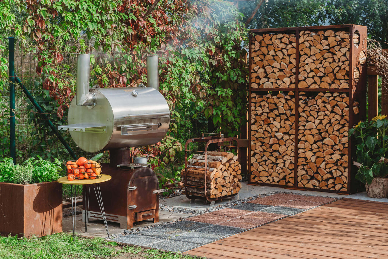 Outdoor wood store crafted from corten steel paired with a large metallic BBQ smoker by Grill Symbol, showcasing a stack of wood within the taller wood store structure. Set against a backdrop of lush greenery and wooden decking with interspersed grass, the image captures the functional and aesthetic synergy between the corten steel wood store and the distinctive BBQ smoker, creating a harmonious outdoor setting
